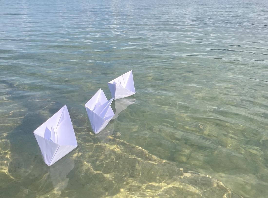 Drei weisse Papierschiffchen schwimmen auf leichten Wellen in seichtem Wasser auf dem Bodensee. Die Sonne spiegelt sich im Wasser und zeichnet Muster auf den Boden. Im Hintergrund sind Segelschiffe auf dem Wasser, dahinter grüne Hügel, und darüber Wolken am Himmel zu sehen.